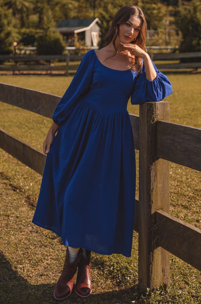 Woman in a blue dress standing next to a wooden fence in a rural setting