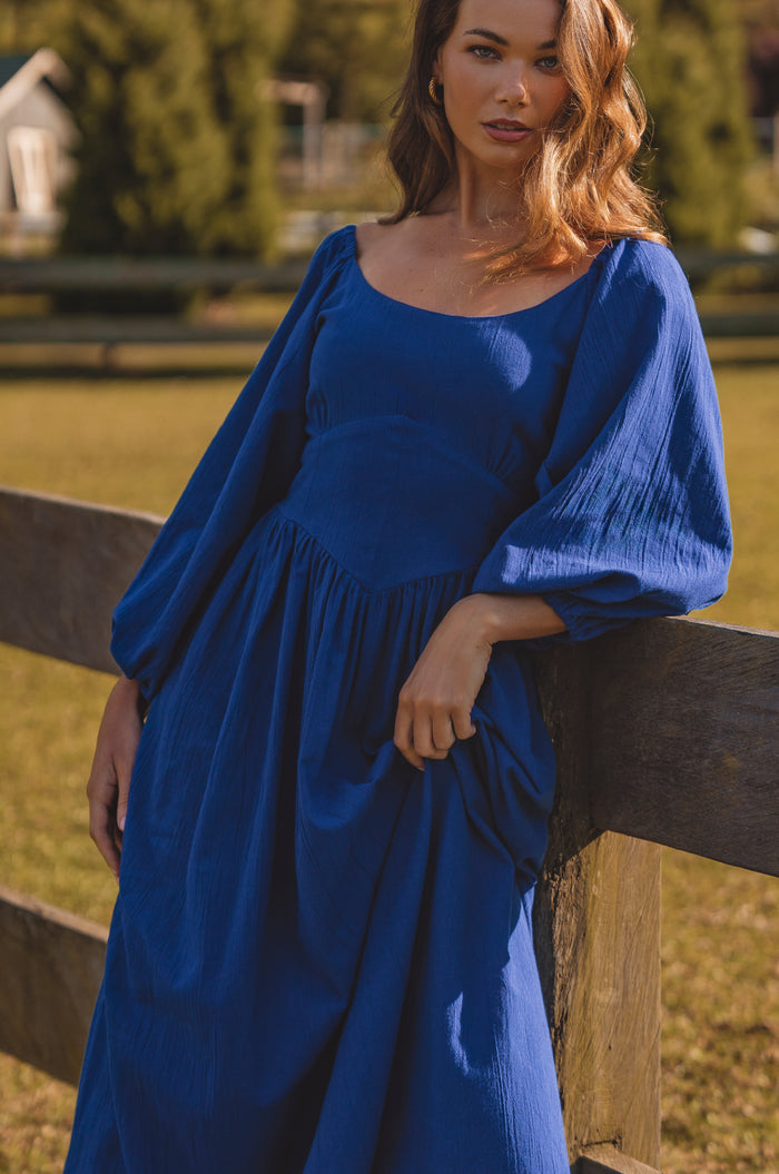 Woman in a blue dress standing by a wooden fence outdoors