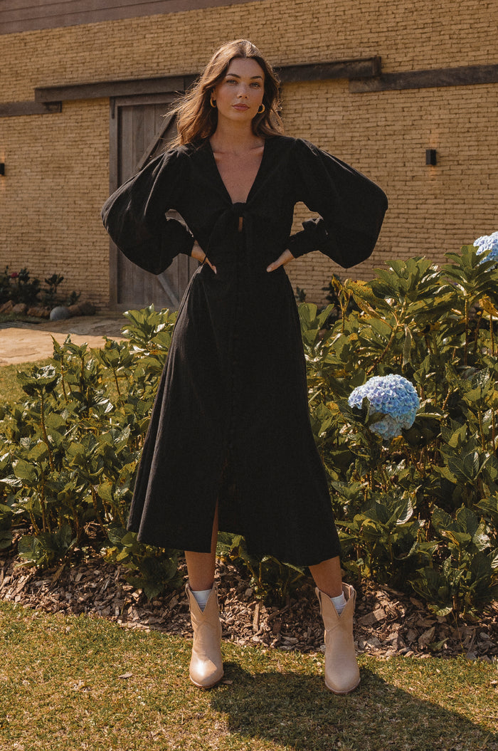 Woman in a black dress standing outdoors with greenery and a building in the background