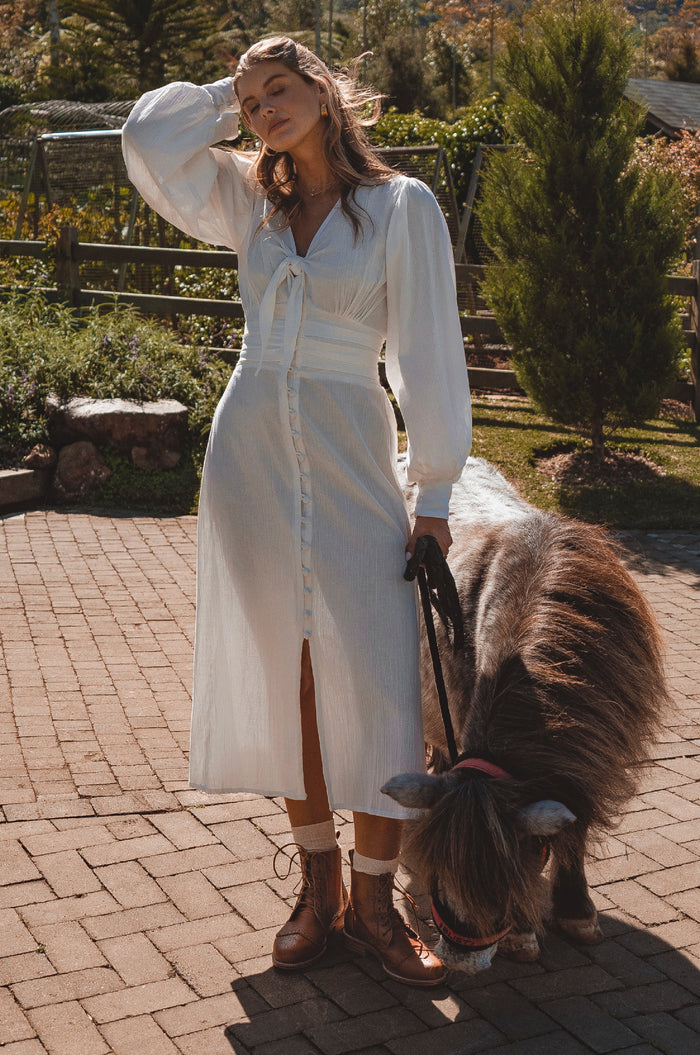Woman in a white dress standing with a small brown pony on a brick path.