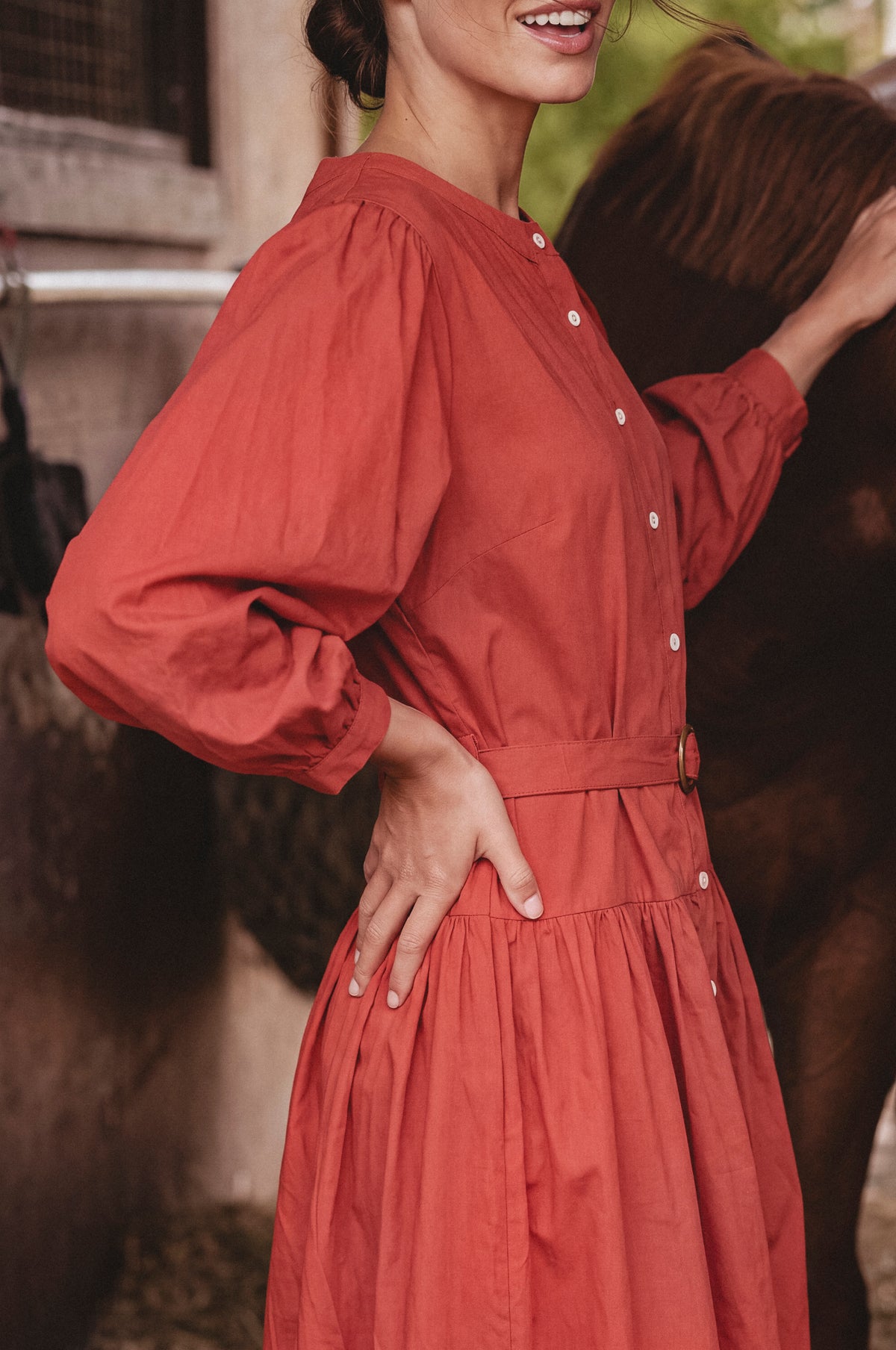 A woman wearing a rust-red long-sleeve button-up dress stands outdoors next to a brown horse, with the dress clearly visible from the chest down.