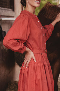 A woman wearing a rust-red long-sleeve button-up dress stands outdoors next to a brown horse, with the dress clearly visible from the chest down.