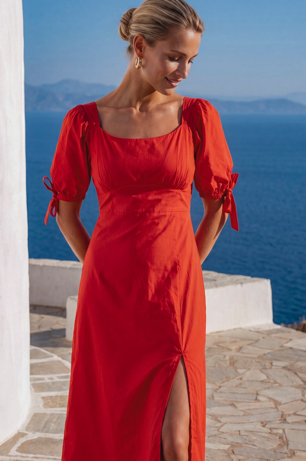 Woman in a red dress standing on a stone path with a blue sea and sky in the background