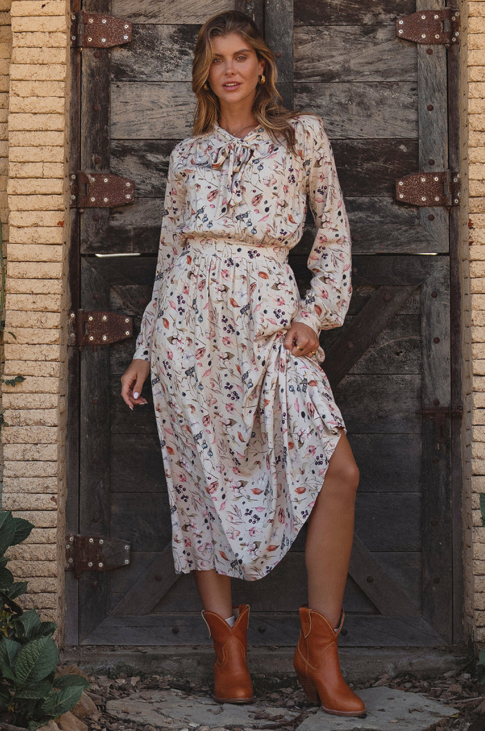 Woman in a floral dress and brown boots standing in front of a rustic wooden door.