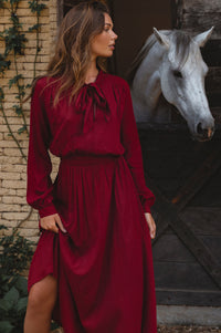 Woman in a red dress standing next to a white horse in front of a wooden door.