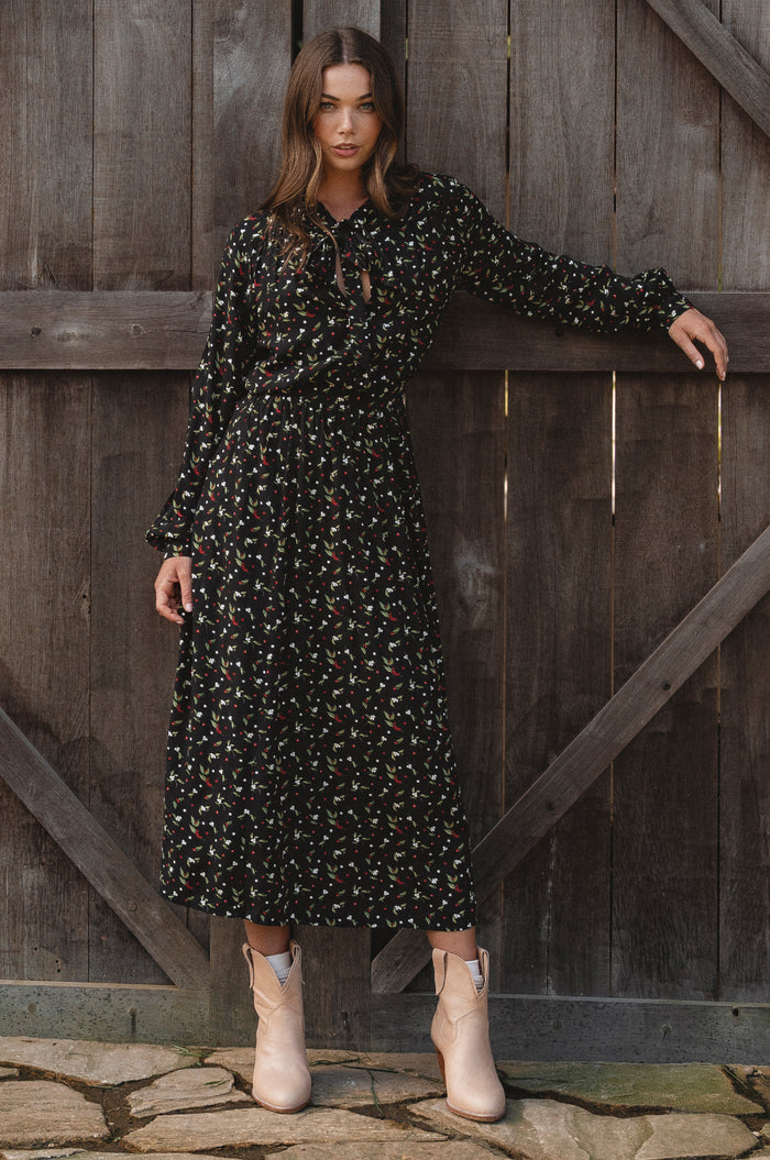 Woman in a black floral dress standing against a wooden gate.