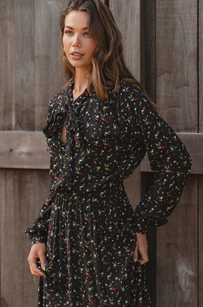 Woman wearing a black floral dress standing against a wooden wall.