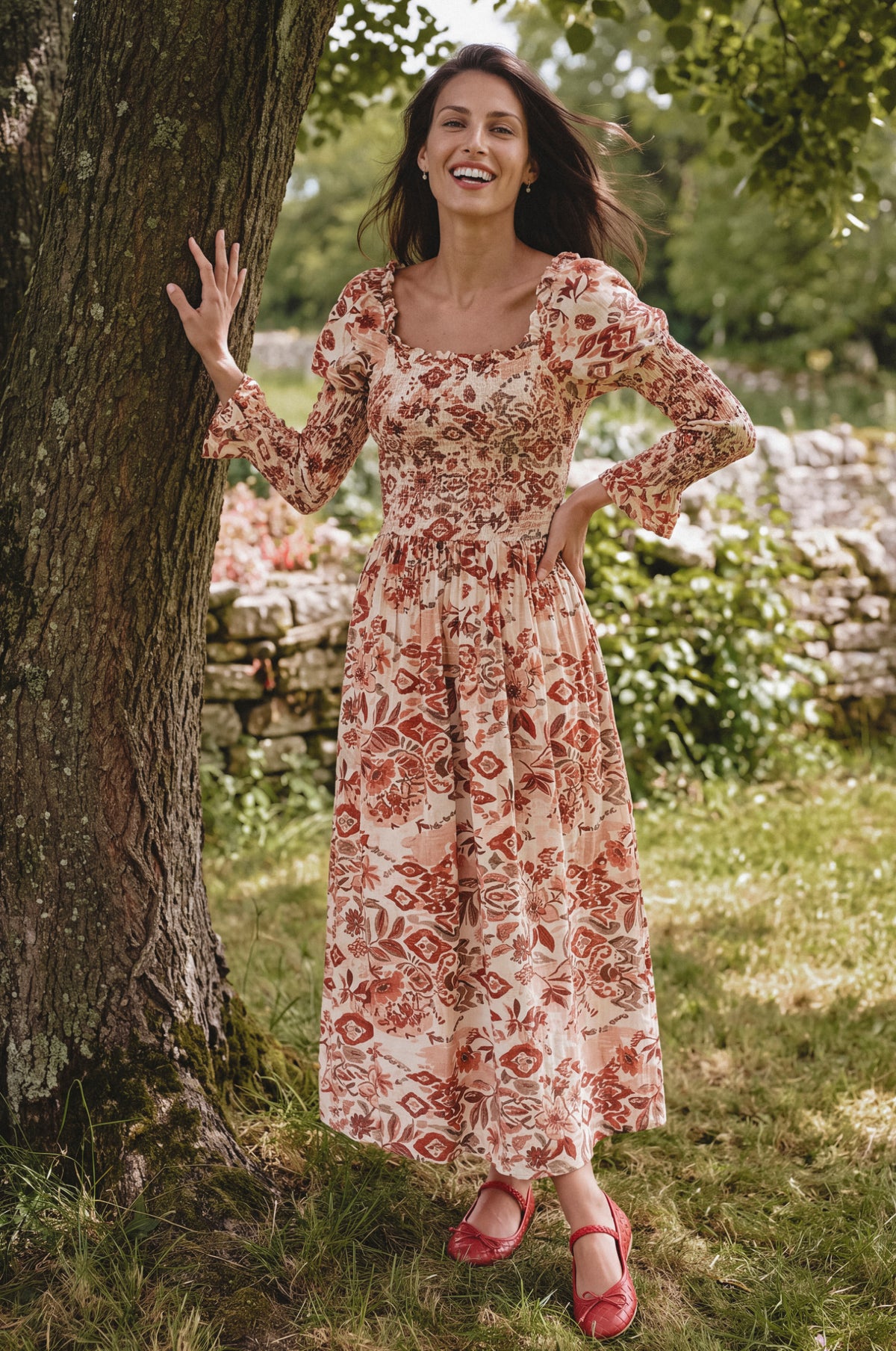 A woman stands outdoors next to a tree wearing a long-sleeved midi dress with a red and orange floral pattern on a cream background, paired with red flat shoes.