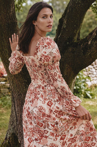 A woman wearing a long-sleeve floral midi dress with a square neckline and puff sleeves, posing outdoors next to a tree trunk.