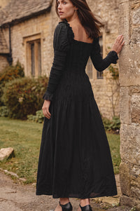 A woman wearing a long black midi dress with long sleeves, a smocked bodice, and a tiered skirt, standing outdoors next to a stone wall in front of a rustic stone building.