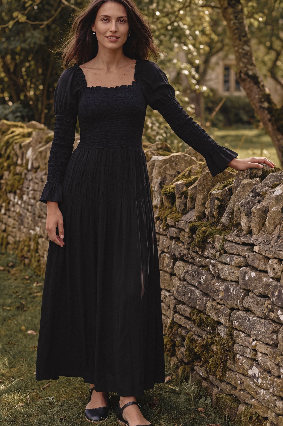 A woman wearing a long black midi dress with puff sleeves and a smocked bodice, standing outdoors next to a mossy stone wall.