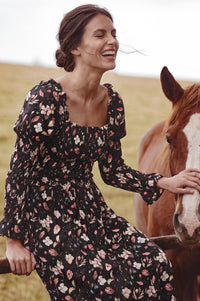 A woman wearing a black floral long-sleeve dress sits beside a brown horse in an outdoor field. The dress has a black base with a multicolor floral pattern in pink, white, and light blue.