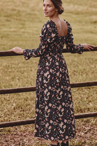 A woman wearing a long-sleeve black midi dress with a pink and beige floral print, standing outdoors in a grassy field next to a wooden fence.