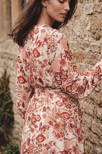 A woman wearing a long-sleeve floral dress with a tie waist, photographed outdoors against a rustic stone wall.