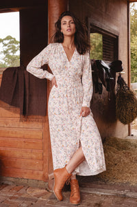 A woman stands in a rustic barn-like setting wearing a long-sleeve white wrap-style maxi dress with a small multicolor floral print and tan ankle boots.