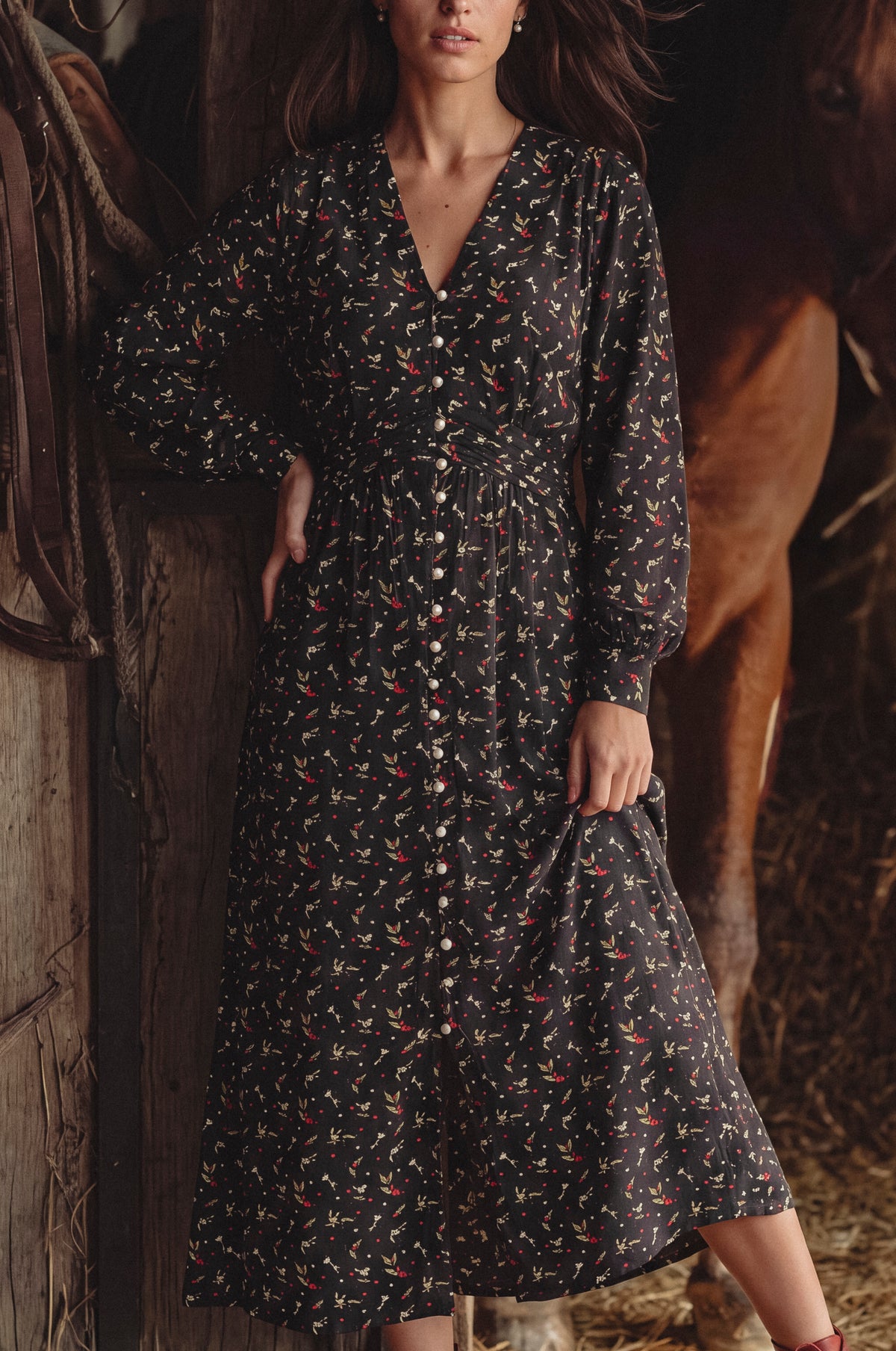 A woman wearing a long-sleeve button-front midi dress with a small multicolor floral print on a dark background, posed in a rustic barn with a horse visible behind her.