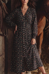 A woman wearing a long-sleeve button-front midi dress with a small multicolor floral print on a dark background, posed in a rustic barn with a horse visible behind her.