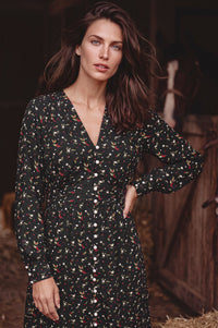 A woman wearing a long-sleeve button-front dress with a small multicolor floral and leaf print on a black background, posed in a rustic barn setting.