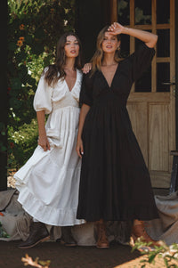 Two women in long dresses standing outdoors with a wooden door in the background.