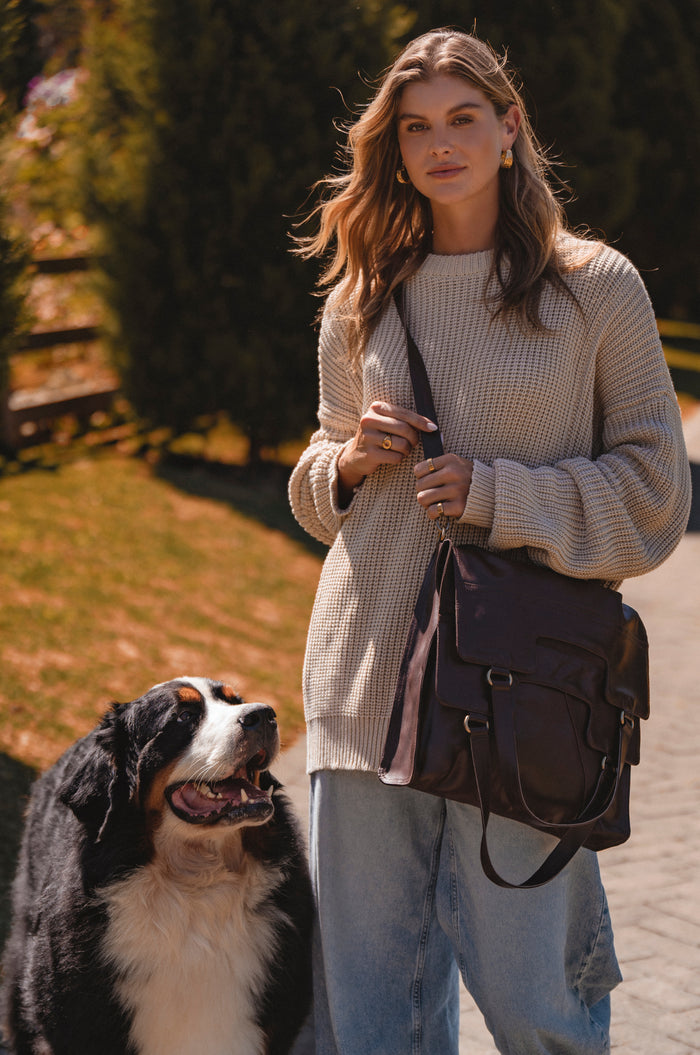 Woman standing outdoors with a dog, holding a brown leather bag.