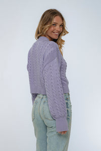 Woman wearing a purple cable knit sweater and light blue jeans on a white background