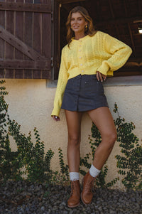 Woman wearing a yellow sweater and dark skirt standing against a rustic wall with wooden door and ivy.