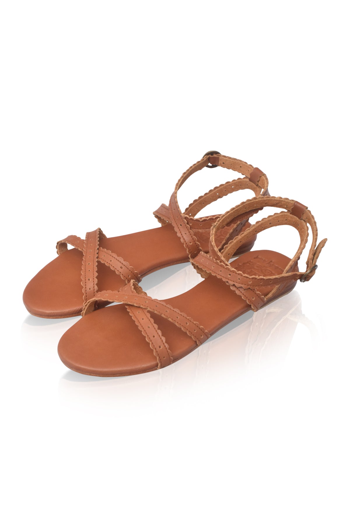 A pair of tan leather strappy flat sandals with adjustable ankle straps, displayed on a clean white background with a soft reflection.