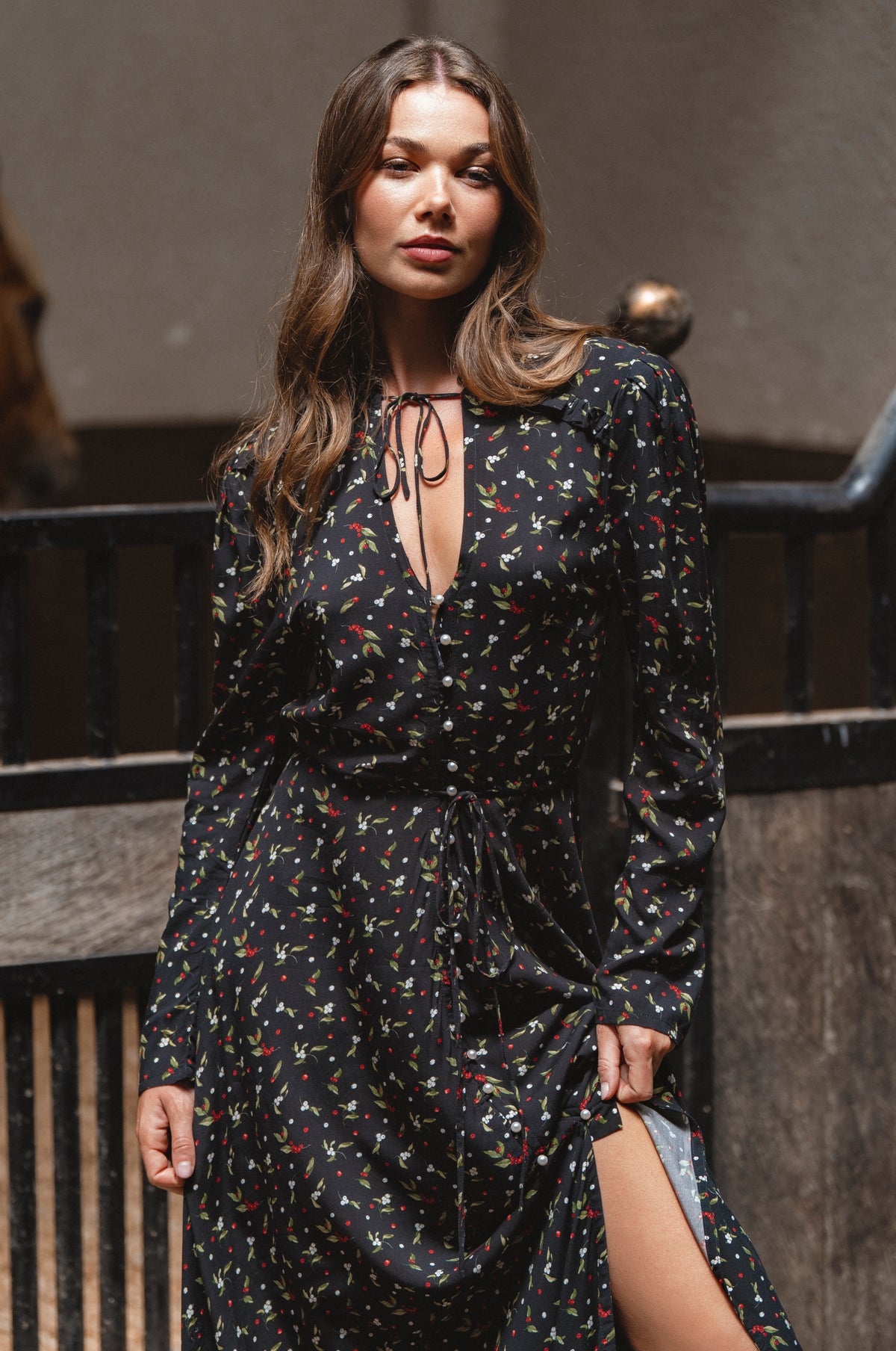 Woman wearing a black floral dress with a slit, standing against a dark background.