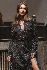 Woman wearing a black floral dress with a slit, standing against a dark background.