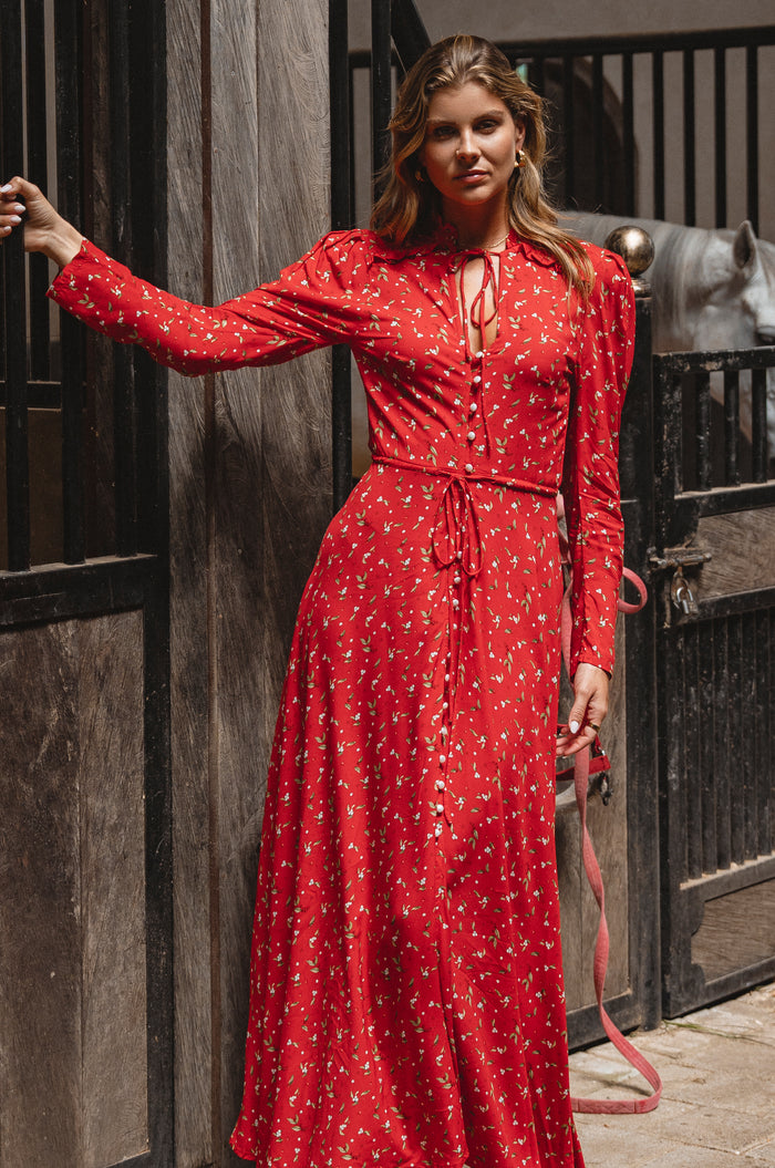 Woman wearing a red dress with a pattern, standing against a wooden wall.
