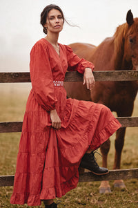 A woman wearing a rust-red tiered maxi dress with long puff sleeves and black lace-up boots leans against a wooden fence in a field, with a brown horse standing behind her.