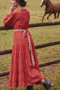 A woman wearing a rust-orange tiered maxi dress with a patterned waist tie, leaning on a wooden fence in a grassy field with a horse in the background.