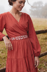 A woman wearing a rust-orange long-sleeve wrap dress with white floral embroidery at the waist, standing outdoors near a wooden fence in a softly blurred field.