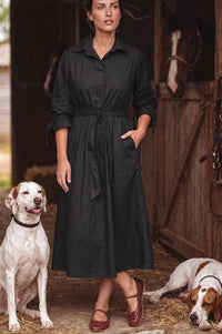 A woman wearing a black long-sleeve midi shirt dress stands in a rustic barn with two dogs on the ground, one sitting white dog on the left and one brown and white dog lying on the right.