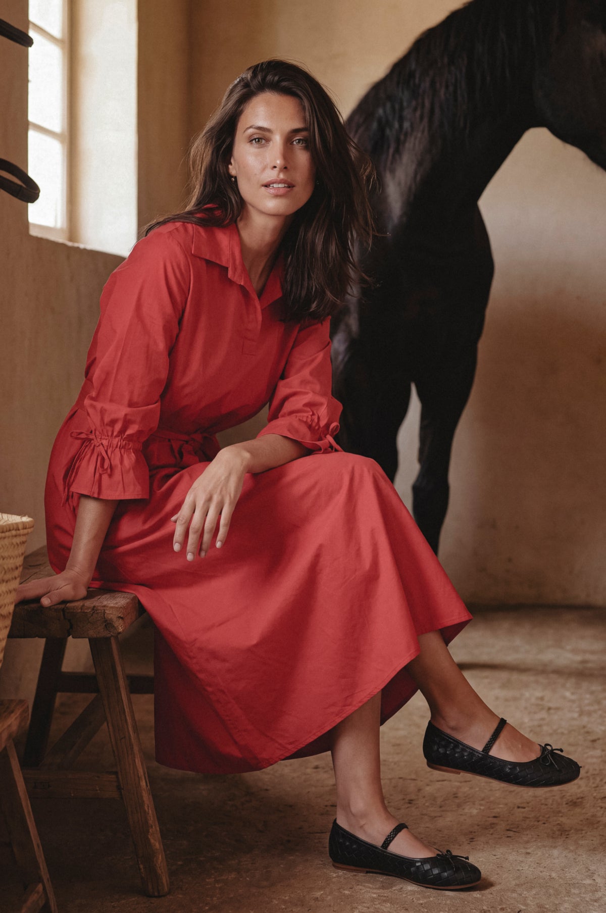 A woman sits on a rustic wooden stool in a warm, earth-toned interior, wearing a long-sleeved red dress and black Mary Jane flats. A dark horse is visible in the background.