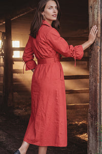 A woman wearing a long-sleeved red midi dress with a tie waist and gathered cuffs, posing in a rustic barn setting.
