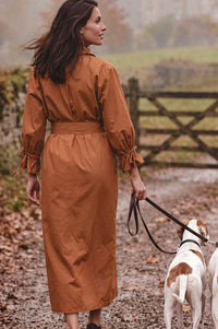 A woman wearing a rust-brown long-sleeve midi dress walks outdoors on a gravel path with a dog on a leash, with a wooden fence and greenery in the background.