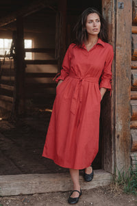 A woman wearing a rust-red midi shirt dress with a tie waist and rolled sleeves, standing in a rustic wooden doorway.