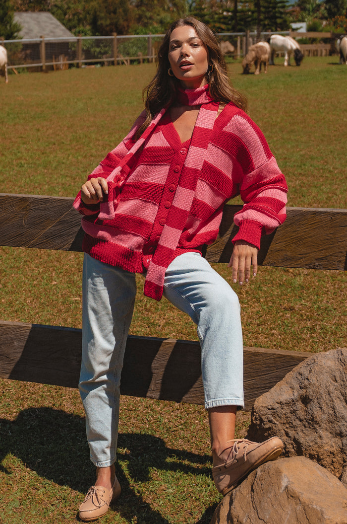 Woman wearing a red and pink checkered cardigan sitting on a wooden fence with a pastoral background.