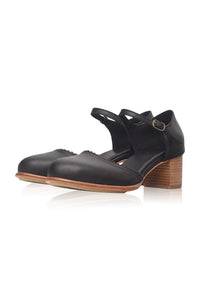 A pair of black leather Mary Jane-style shoes with a low block heel, adjustable ankle straps, and a small gold buckle, shown on a white background with a soft reflection.