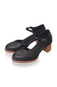 A pair of black leather Mary Jane style shoes with low block heels, ankle straps, and a small gold buckle, shown on a white background with a soft reflection.