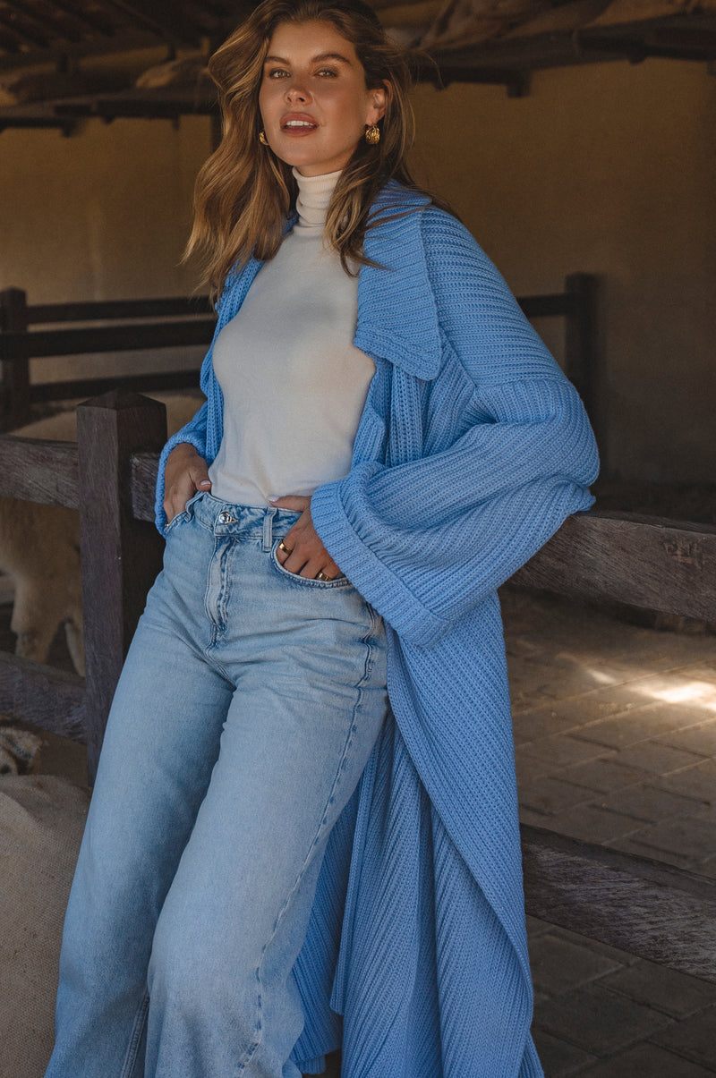 Woman wearing a blue knitted cardigan over a white top and jeans, standing in a rustic indoor setting.